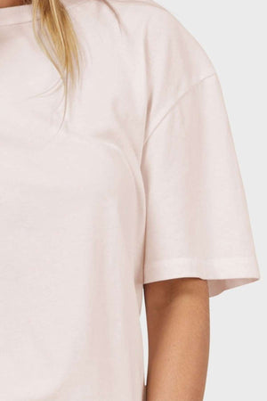A person in a beige Academy Women Jimmy Boyfriend Tee, crafted from premium sanded jersey. The image focuses on the upper half and shoulder of this oversized short-sleeved T-shirt, set against a minimal background.