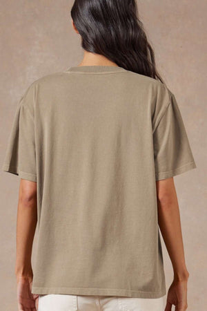 A person with long dark hair wears the Academy Women Jimmy Boyfriend Tee, an oversized premium sanded jersey top, paired with light-colored pants and stands facing away from the camera against a neutral background.