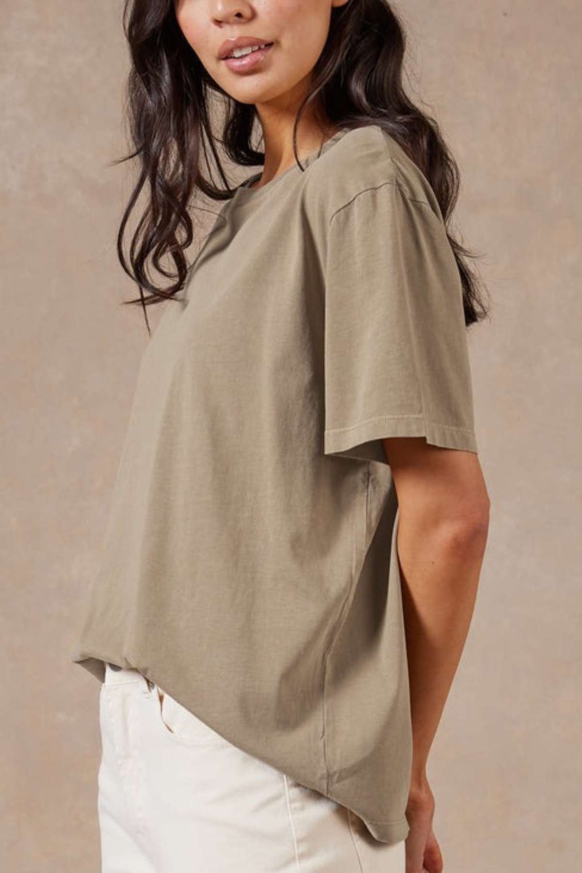 A woman with long dark hair wears the Academy Women Jimmy Boyfriend Tee, an oversized sanded jersey top, paired with off-white pants. She stands against a neutral background, shown from the waist up and slightly turned to the side.
