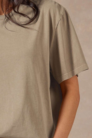 A person with long, dark hair wears the Academy Women Jimmy Boyfriend Tee—a loose, short-sleeved beige top made from premium sanded jersey—shown from shoulders to waist against a neutral background.