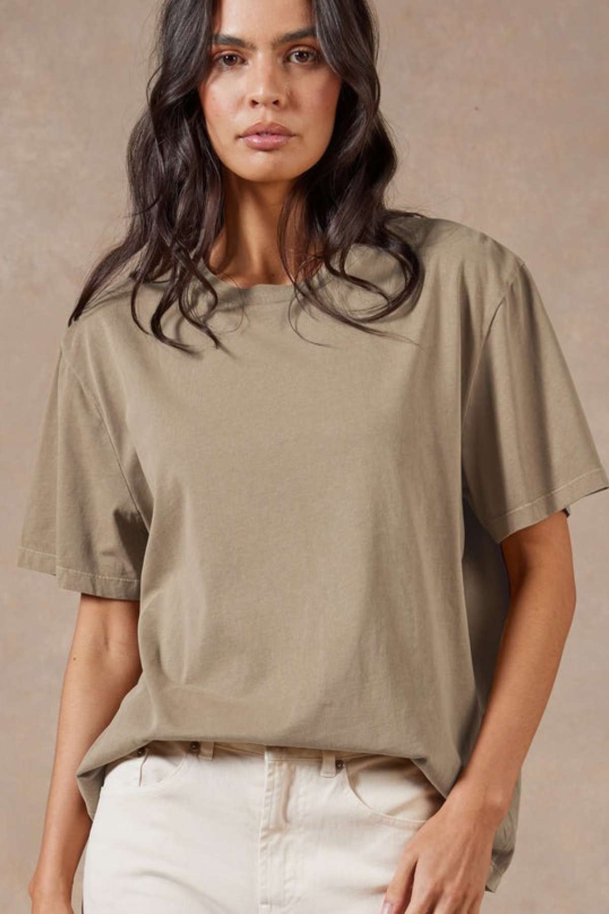 A woman with long dark wavy hair wears the Academy Women Jimmy Boyfriend Tee in premium sanded jersey and light pants, standing against a plain, neutral background.