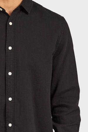 A person wearing the Academy Brand Harry Double Cloth Shirt, a long-sleeve black button-up with white buttons, shown from shoulders to mid-torso against a plain light background.