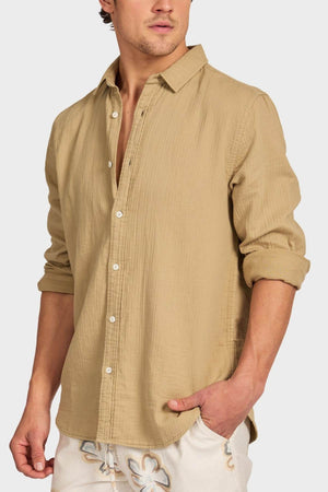 A man wears the Academy Brand Harry Double Cloth Shirt—a light brown, casual cotton shirt with rolled sleeves and one hand in his pocket—paired effortlessly with white floral-patterned shorts.