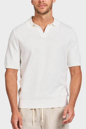 A man wears the Academy Brand Jonny Textured Knit Polo in white, featuring a collar and open neckline, paired with light beige drawstring pants, standing against a plain light background.