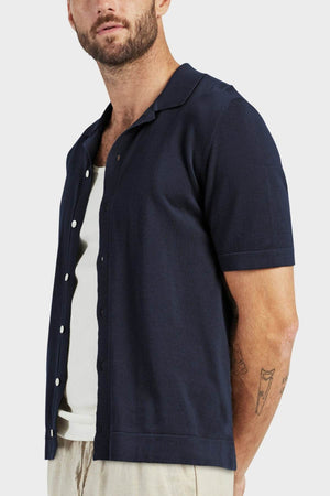A bearded man with a tattoo on his left forearm wears the Academy Brand Jackson Knit Short Sleeve Shirt over a white tank top and beige pants, standing against a plain light background.
