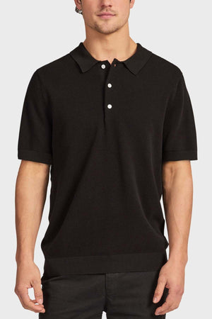 A man stands against a plain, light background wearing the Academy Brand Riley Knit Polo in black, featuring short sleeves, a collar, and three white buttons. The breathable fabric provides comfort as his arms rest relaxed by his sides.