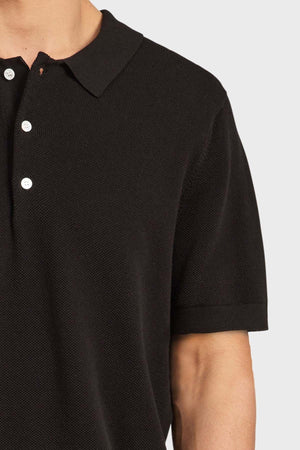 A person wears the Academy Brand Riley Knit Polo, a textured, breathable polo with a collar and three white buttons, shown from shoulders to just above the elbow against a plain light background.