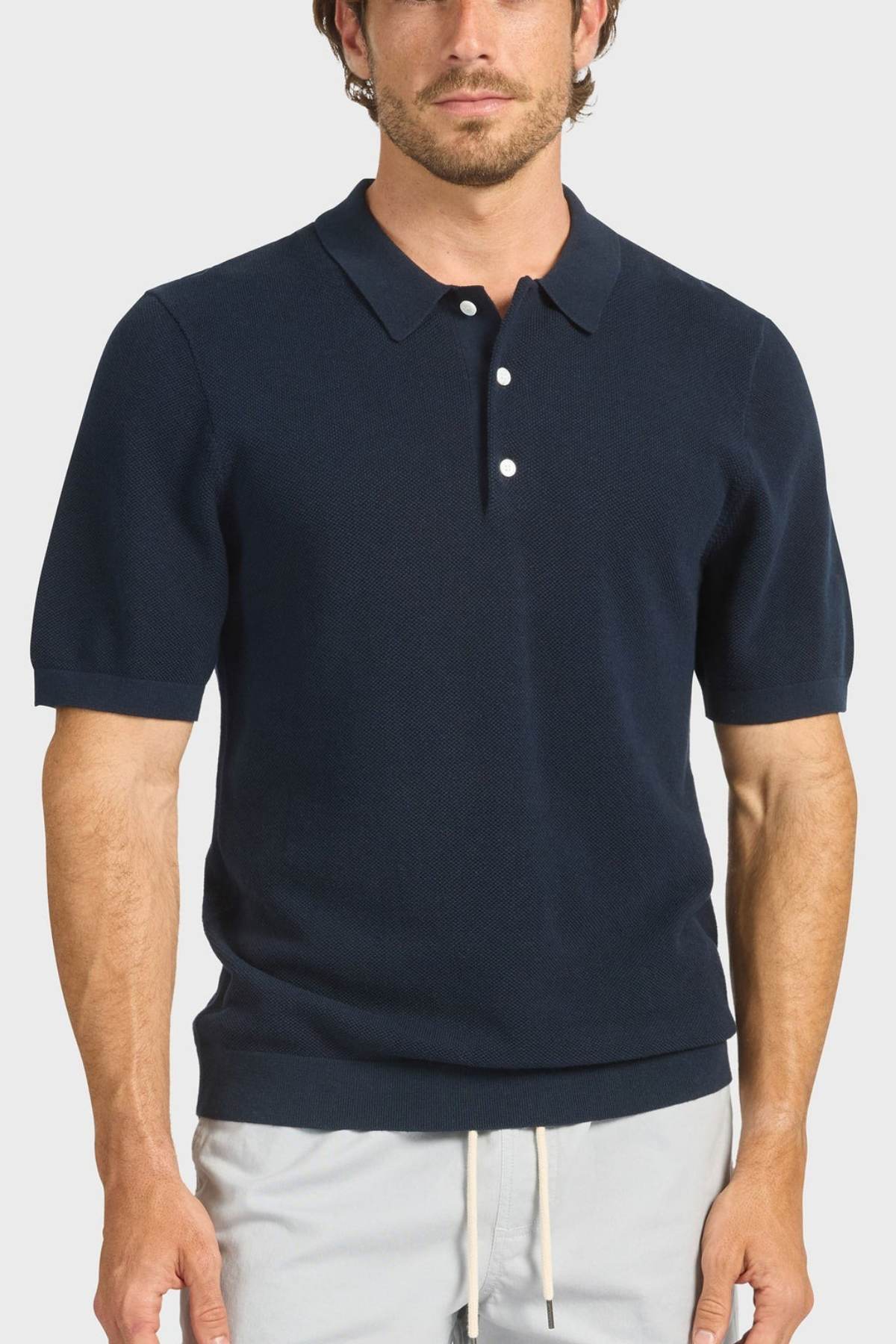 A man wears the Academy Brand Riley Knit Polo in navy blue with white buttons and breathable fabric, paired with light drawstring pants, standing against a plain light background.
