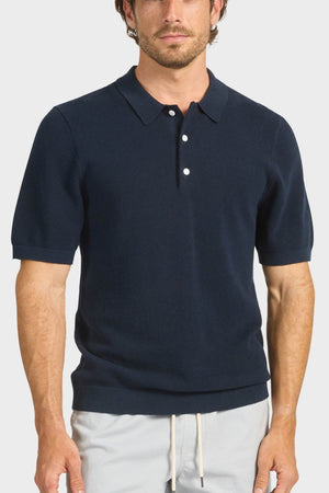 A man wears the Academy Brand Riley Knit Polo in navy blue with white buttons and breathable fabric, paired with light drawstring pants, standing against a plain light background.