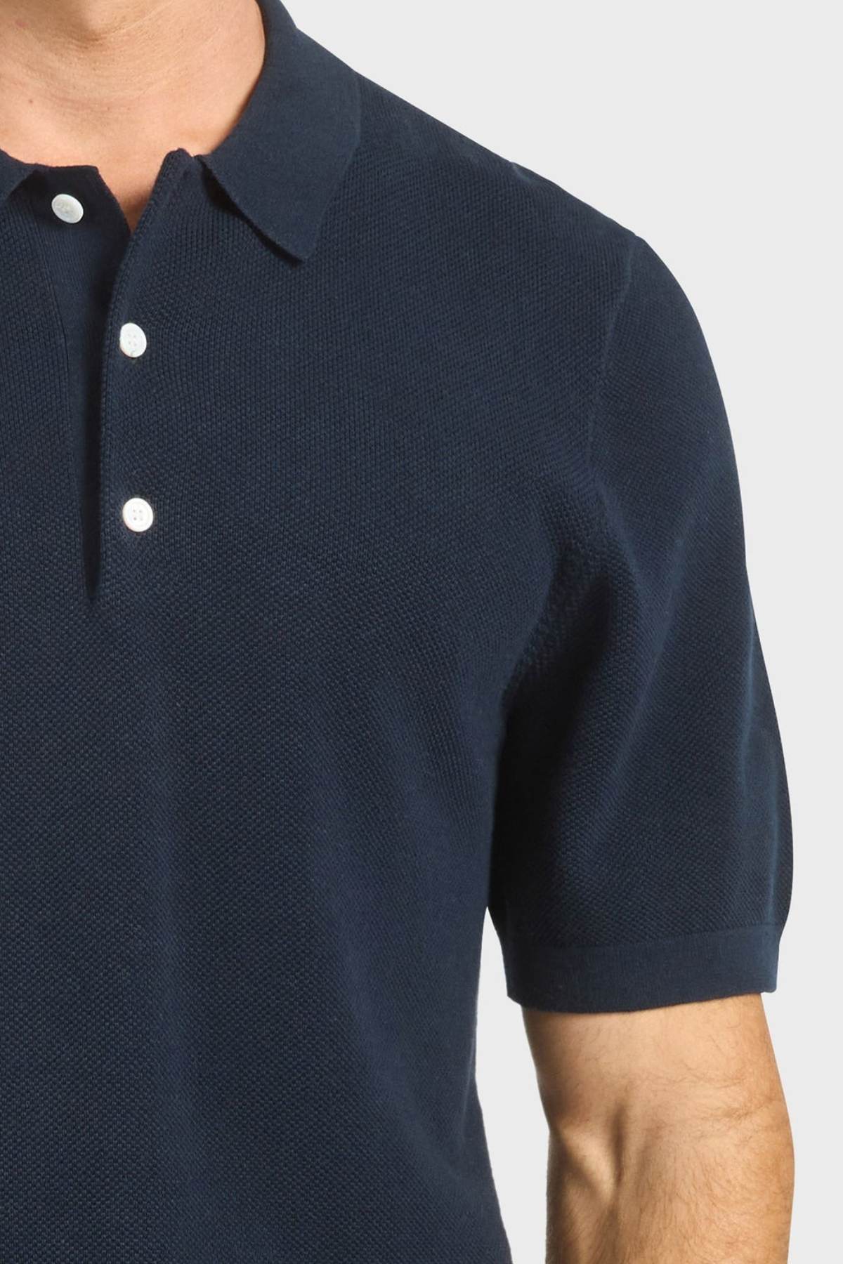 A person wears the Academy Brand Riley Knit Polo in navy blue with white buttons, shown from shoulders to mid-torso against a plain light background. The breathable fabric ensures comfort in this classic short-sleeve style.