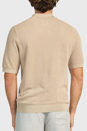 A man is shown from the back wearing an Academy Brand Riley Knit Polo in light beige with short sleeves and light gray pants. The textured stitch of the polo stands out against a plain white background.