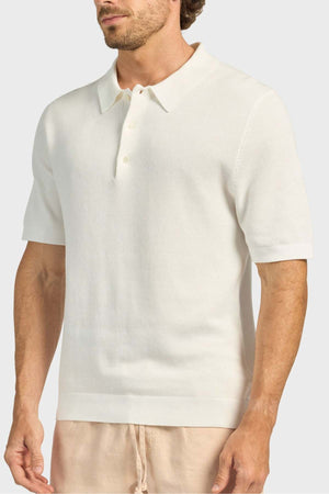 A man wears the Academy Brand Riley Knit Polo, a short-sleeve off-white textured polo made from breathable fabric, paired with beige pants, standing against a plain light gray background.