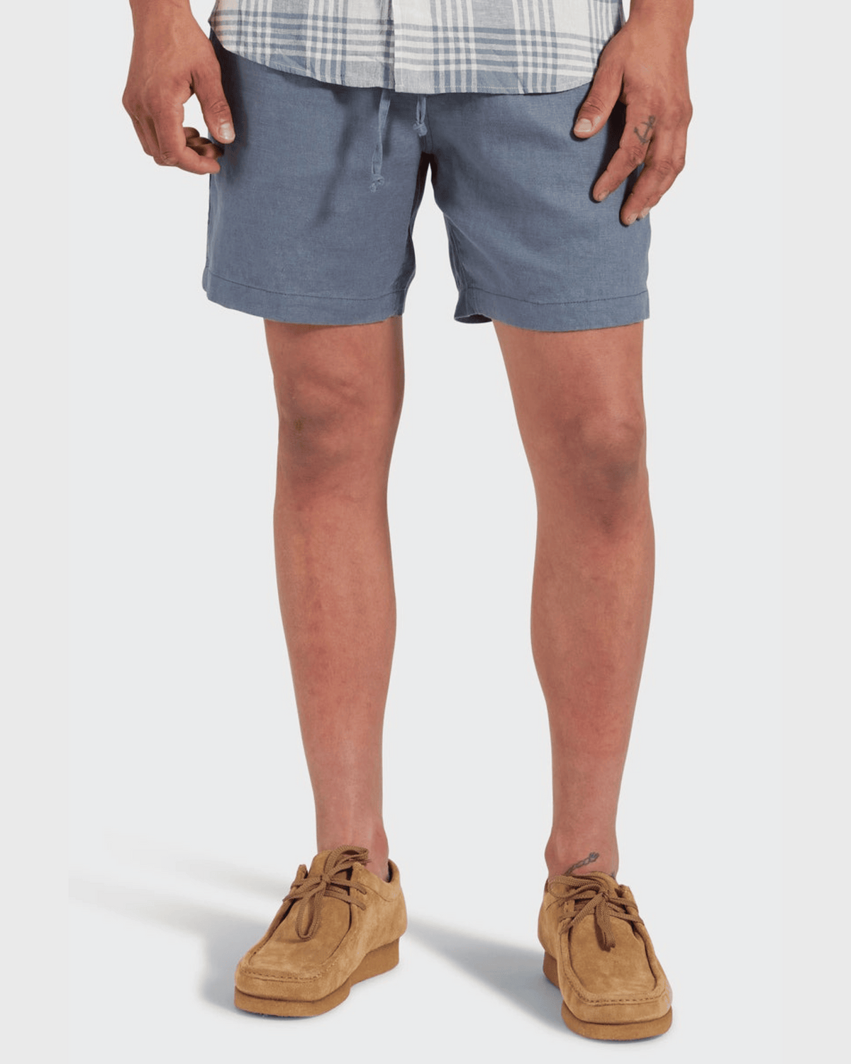 A person stands from mid-torso to feet against a plain white background, wearing Academy Brand Riviera Linen Shorts in relaxed fit light blue with an elastic waist, paired with a gray and white plaid shirt and tan suede shoes.