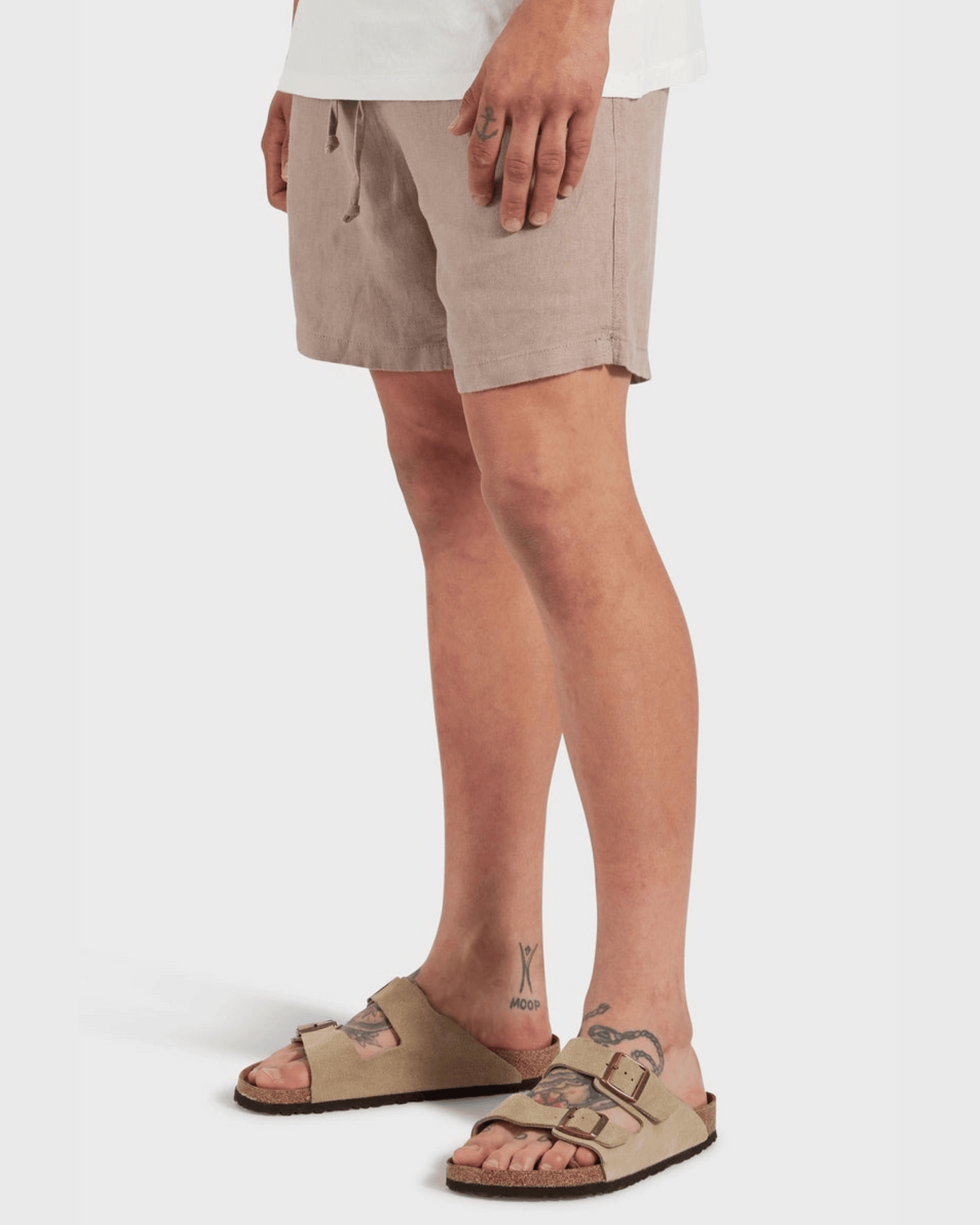 A person wearing Academy Brand Riviera Linen Short in light brown with an elastic waist, a white shirt, and beige two-strap sandals stands on a white background. Tattoos are visible on their legs.