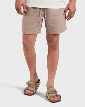 A person wearing Academy Brand Riviera Linen Shorts in light brown, a white shirt, and tan buckle sandals stands against a plain white background. Tattoos are visible on their left hand and both legs.