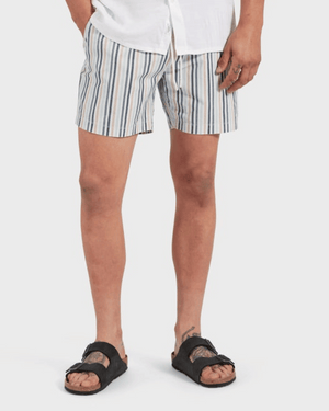 Wearing an Academy Brand Teamster Stripe Boardy, a person in a white shirt and black sandals stands with tattoos on one leg and hand. The background's simple white sets off the vintage vibe of their blue-and-white striped shorts made from recycled polyester.
