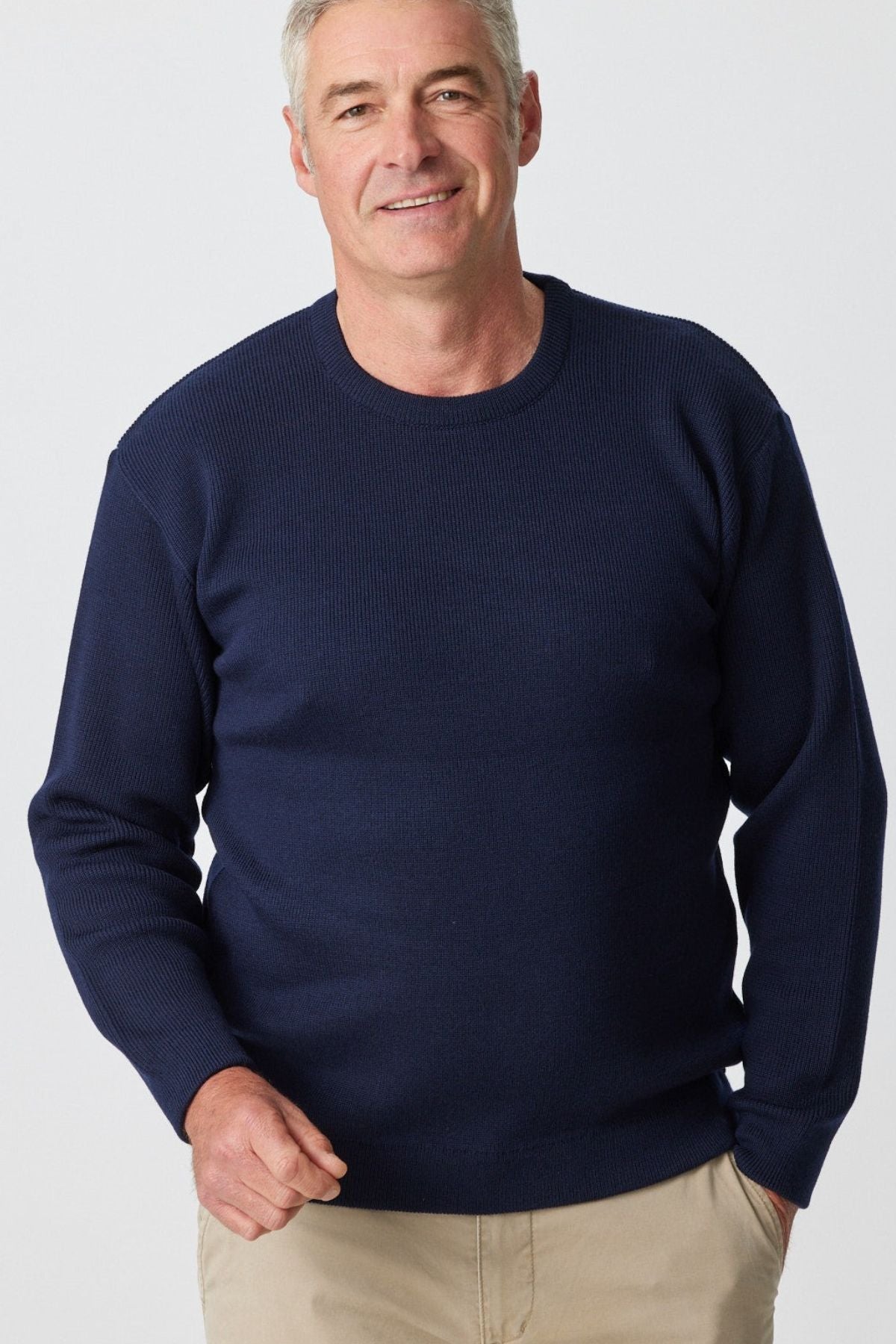 A person wearing an Aklanda Fishermans Rib Crew Pullover in dark blue and beige pants stands against a plain background, smiling at the camera.