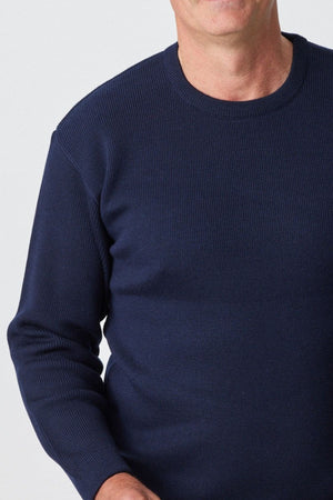 A person wearing the Aklanda Fishermans Rib Crew Pullover in navy blue stands against a plain white background, visible from the shoulders to the waist. The person's face is not visible in the image.