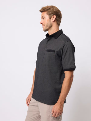 A man with light brown hair models the Breakaway Rafael Micro Prima Polo, featuring a black pocket detail and made from a cotton-polyester blend, paired with light pants while standing sideways against a plain, light background.