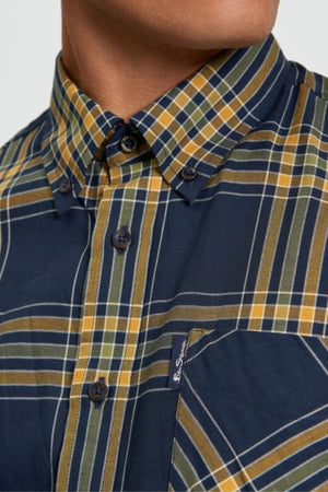 A close-up of a person wearing the Ben Sherman Grid Check Shirt, featuring a buttoned-up navy blue base with yellow, green, and white plaid, plus a button-down collar and front pocket.