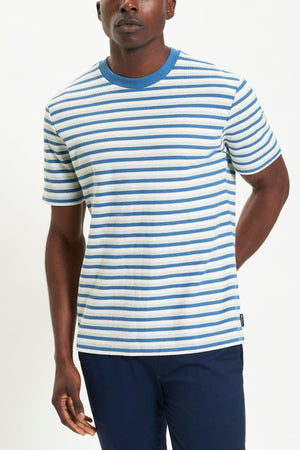 A person stands against a plain white background wearing the Ben Sherman Honeycomb Stripe Tee—short-sleeved with white and blue horizontal stripes and a blue collar—paired with dark blue pants.