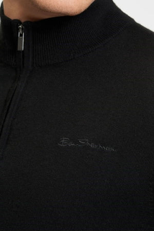 Close-up of a person wearing the Ben Sherman Merino Half Zip Knit in black, featuring the embroidered "Ben Sherman" logo on the chest. Only their lower face and upper torso are shown.