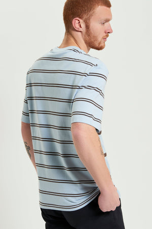 A man with short red hair and a beard stands sideways, wearing a relaxed fit Ben Sherman Mixed Stripe Tee with black and white stripes, black pants, and a tattoo on his left arm. His left hand is in his pocket.