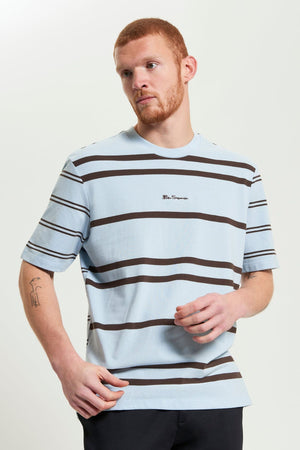 A man with short red hair and a beard wears the Ben Sherman Mixed Stripe Tee—light blue pique cotton with black and white horizontal stripes, a small center logo, and a relaxed fit—standing against a plain light background.