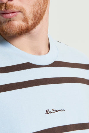 A man with a reddish beard wears the Ben Sherman Mixed Stripe Tee in light blue pique cotton with brown stripes and “Ben Sherman” embroidered on the chest, against a plain background. His face is partially visible.