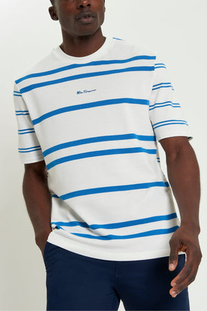Wearing the Ben Sherman Mixed Stripe Tee in white and blue with navy pants, this person stands confidently with one hand in their pocket against a plain background.