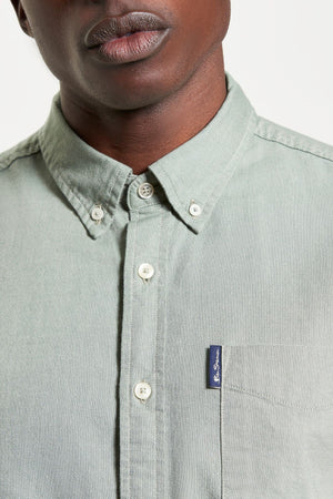 A close-up of a person wearing the Ben Sherman Signature Oxford Short Sleeve Shirt in light green, featuring a chest pocket with a blue tag and button-down collar. Only the lower half of the face is visible.