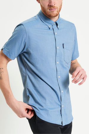 A man with a short beard wears a light blue Ben Sherman Signature Oxford Short Sleeve Shirt and black pants, standing against a plain white background. His right hand is at his side and his left hand is slightly raised.
