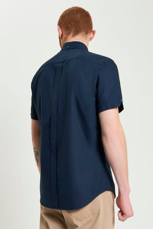 A short-haired person with red hair is seen from behind in a navy Ben Sherman Signature Oxford Short Sleeve Shirt made of organic cotton, featuring a straight back seam, collar button, and paired with beige pants.