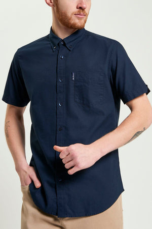 A man with a trimmed beard wears a navy Ben Sherman Signature Oxford Short Sleeve Shirt, made from organic cotton with a chest pocket, paired with beige pants. He stands with one hand in his pocket and holds the shirt with the other.