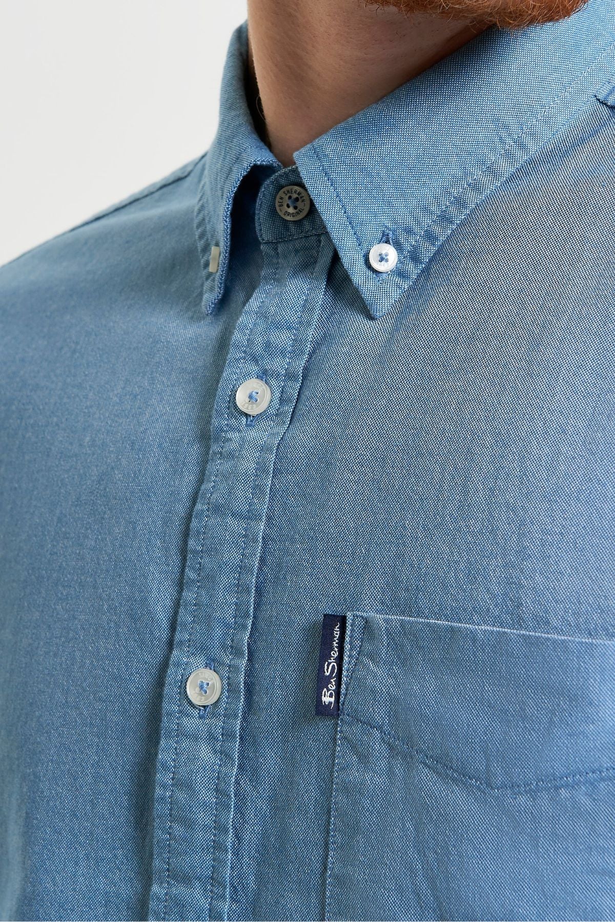 Close-up of a person wearing a light blue Ben Sherman Signature Oxford Short Sleeve Shirt made from organic cotton, with a collar, white buttons, and a small navy "Ben Sherman" tag on the pocket. Only the lower face, neck, and upper chest are shown.