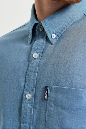 Close-up of a person wearing a light blue Ben Sherman Signature Oxford Short Sleeve Shirt made from organic cotton, with a collar, white buttons, and a small navy "Ben Sherman" tag on the pocket. Only the lower face, neck, and upper chest are shown.