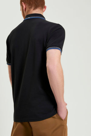 A man with short hair is seen from behind, wearing a Ben Sherman Signature Pique Polo with blue and white trim on the collar and sleeves, paired with light brown pants. One hand is in his pocket against a plain light background.