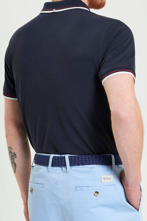 A man facing away wears a Ben Sherman Signature Pique Polo with white and red trim, light blue belted shorts, and displays a tattoo on his left arm against a plain, light background.
