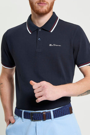 A man wears a Ben Sherman Signature Pique Polo with white and red trim. Crafted from soft organic cotton, the shirt features a small logo on the chest. Only his upper body is visible, paired with light blue pants and a woven blue belt.