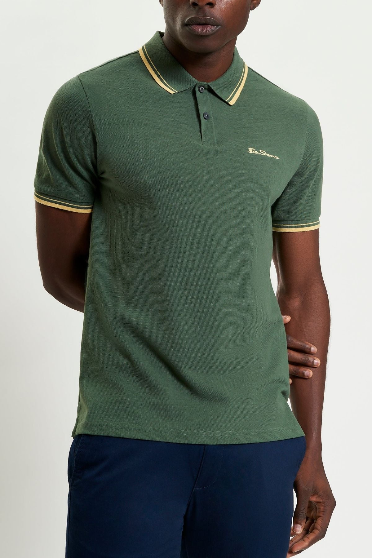 A man wears a Ben Sherman Signature Pique Polo in green with yellow trim on the collar and sleeves, paired with dark blue pants. He stands against a plain background, with one arm at his side and the other behind his back.
