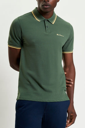 A man wears a Ben Sherman Signature Pique Polo in green with yellow trim on the collar and sleeves, paired with dark blue pants. He stands against a plain background, with one arm at his side and the other behind his back.