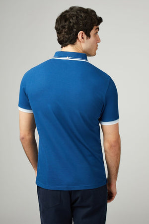 A man with short curly hair stands with his back to the camera, wearing a Ben Sherman Signature Pique Polo in blue with white trim on the collar and sleeves, paired with dark pants. The background is plain and light-colored.