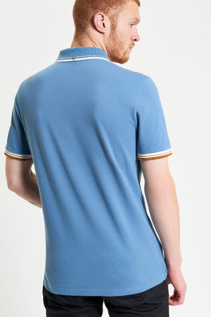 A man with short red hair and a beard is shown from the back, wearing a light blue Ben Sherman Signature Pique Polo with white and brown sleeve trim and black pants, set against a plain white background.