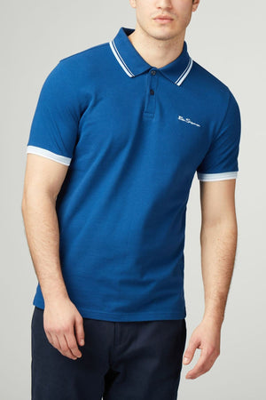 A man stands against a plain light background wearing the Ben Sherman Signature Pique Polo in blue with white trim on the collar and sleeves, paired with dark trousers.
