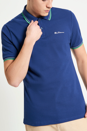 A person wears a Ben Sherman Signature Pique Polo in blue with green and white trim on the collar and sleeves, paired with beige pants. The shirt has a small embroidered logo on the left chest. The personβs head is partially out of frame.