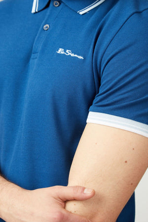 A person is shown from shoulders to forearm wearing the Ben Sherman Signature Pique Polo in blue with white trim, crafted from soft organic cotton, holding their left elbow with their right hand.