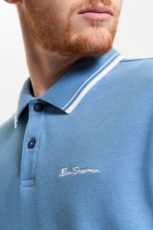 Close-up of a man with a reddish beard wearing the Ben Sherman Signature Pique Polo in regular fit, blue with white collar trim and "Ben Sherman" embroidered on the chest. The man’s face is partially visible.