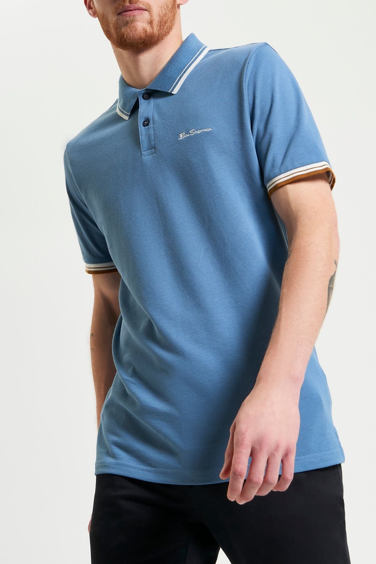 A man wears a blue Ben Sherman Signature Pique Polo with white and yellow striped collar and cuffs in regular fit, paired with black pants, standing against a plain light background.