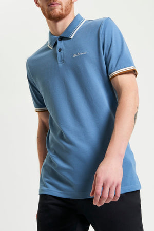 A man wears a blue Ben Sherman Signature Pique Polo with white and yellow striped collar and cuffs in regular fit, paired with black pants, standing against a plain light background.