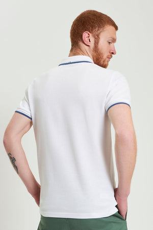 A man with short red hair and a beard, tattoo visible on his left arm, stands with his back to the camera wearing green pants and a white Ben Sherman Signature Pique Polo with blue trim, made from organic cotton.